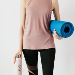 Faceless slim female athlete in sportswear standing with blue fitness mat and water bottle while preparing for indoors workout